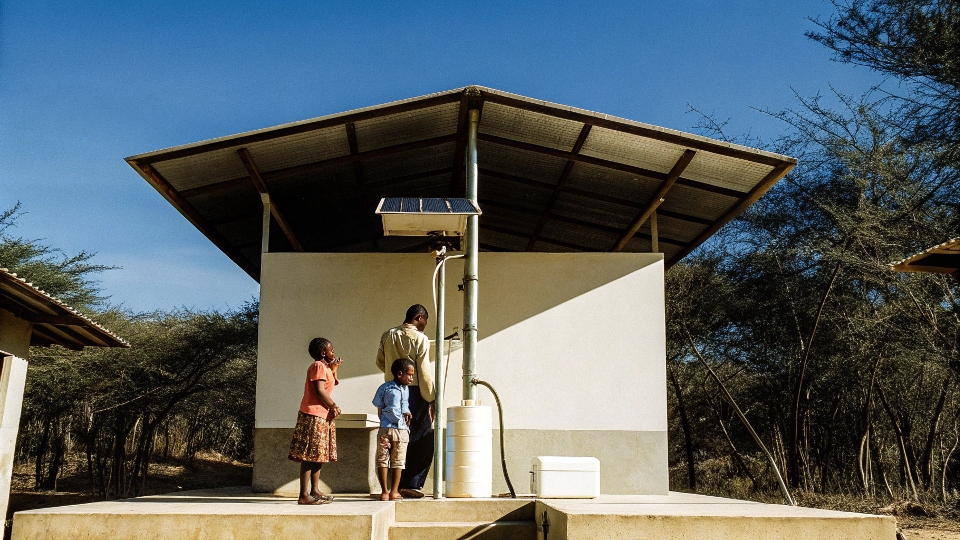 A community water kiosk in an African village with people filling jerrycans from taps, with a solar panel array nearby